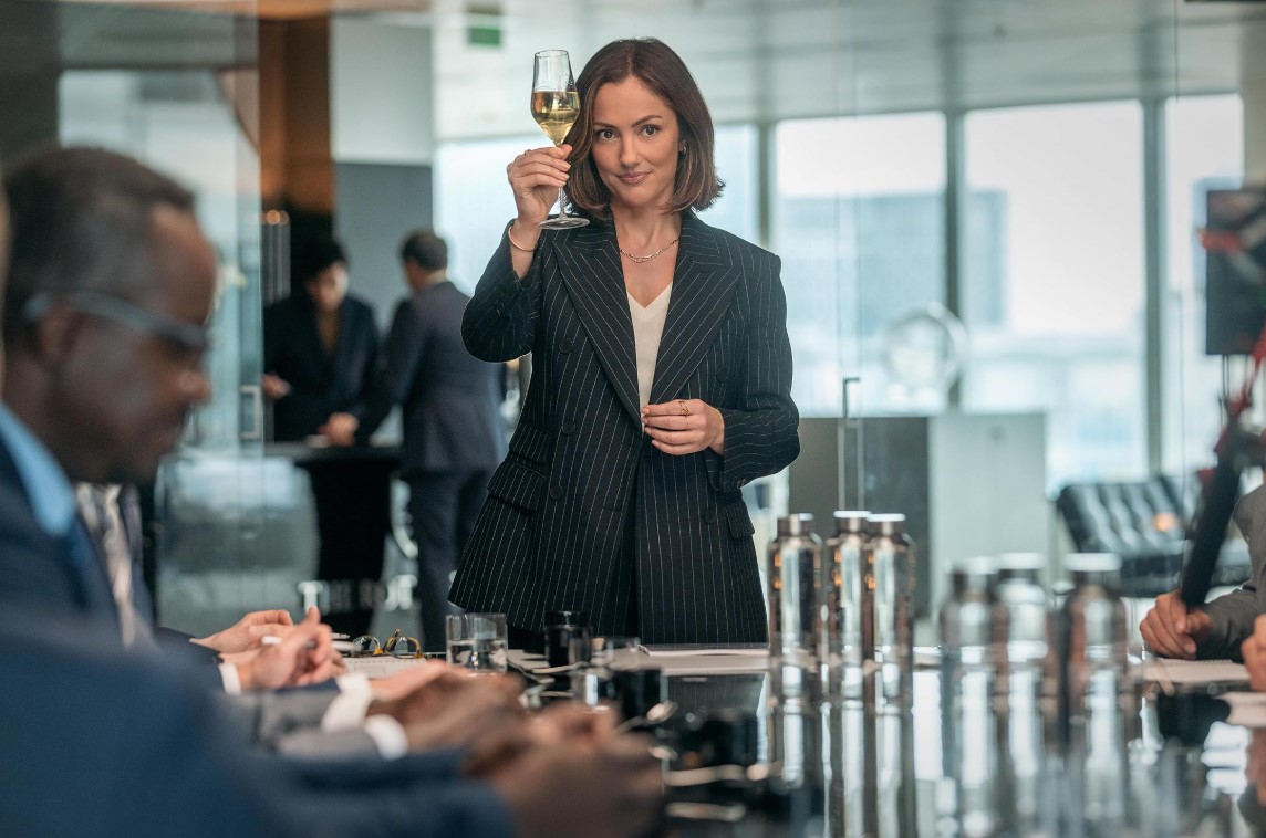 샴페인 프라블럼에서 시드니 프라이스 역의 민카 켈리가 회의실에서 샴페인 잔을 들고 있는 장면 – M&A 임원으로서의 강인한 모습을 보여주는 스틸컷