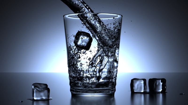 푸른 배경 앞에서 얼음이 담긴 유리컵에 시원한 물이 빠르게 붓고 있으며, 물방울이 튀는 생생한 장면이 포착된 이미지. 수분 섭취의 중요성과 관련된 내용을 시각적으로 강조하는 사진입니다.