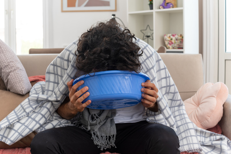 sick-young-caucasian-man-optical-glasses-wrapped-plaid-with-scarf-around-his-neck-holding-vomiting-into-bucket-sitting-couch-living-room-900
