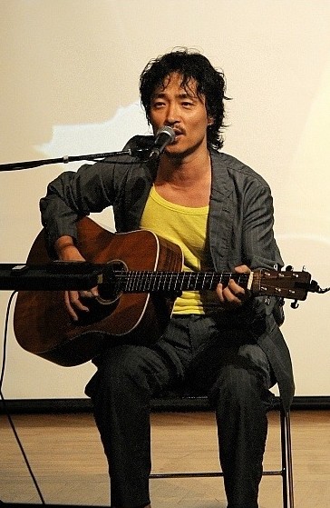 거꾸로-강을-거슬러-오르는-저-힘찬-연어들처럼-강산에가-노란-티셔츠에-빈티지한-회색-슈트를-입고-의자에-앉아-기타를-치며-노래하고-있는-사진