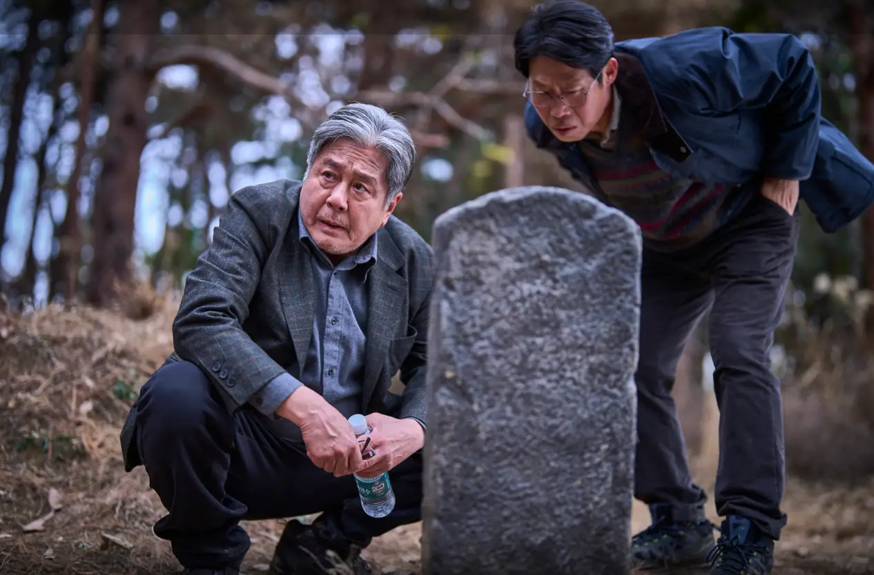 파묘-양복입은 최민식이 비석앞에 쪼그리고 앉아 있고 그옆 유해진이 엎드려 비석을 바라보고 있는 모습