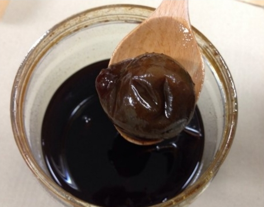 야생에서 채취를 한 매실을 풀 고와서 만든 검은 매실청을 투명하고 둥근 유리 그릇에 반쯤 담아두고 나무숟가락으로 삶은 매실 한 개를 담아서 유리그릇 위에 걸쳐놓고 직은 사진