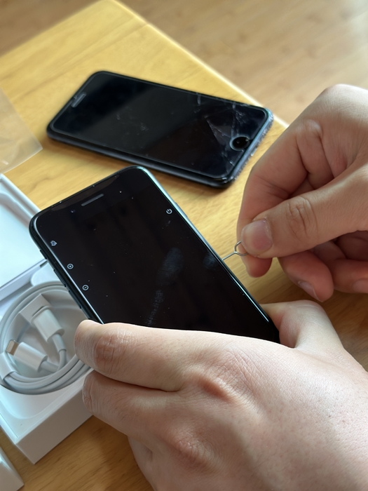 유심 핀으로 아이폰 SE3 측면 유심 트레이를 열고 있는 모습&amp;#44; 뒤쪽에는 액정이 깨진 아이폰 7이 함께 놓여 있음