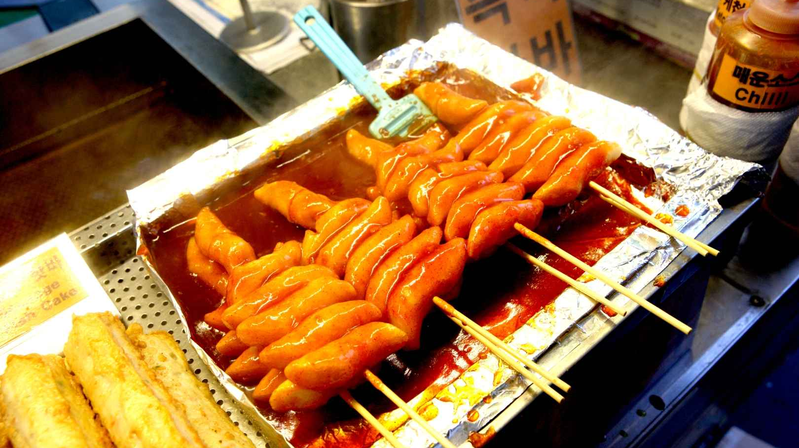 Korean street food tteokkochi skewers being glazed with sweet and spicy gochujang sauce at a market stall