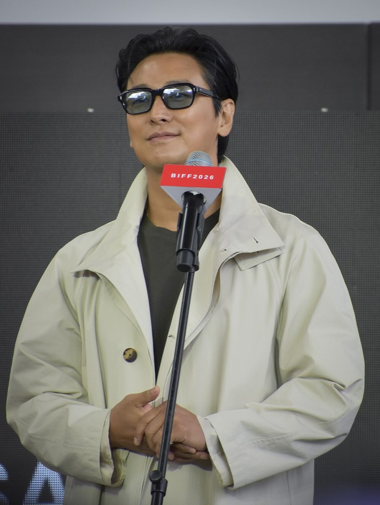 선글라스를 쓰고 밝은 색 코트를 입은 채 마이크 앞에 선 주지훈