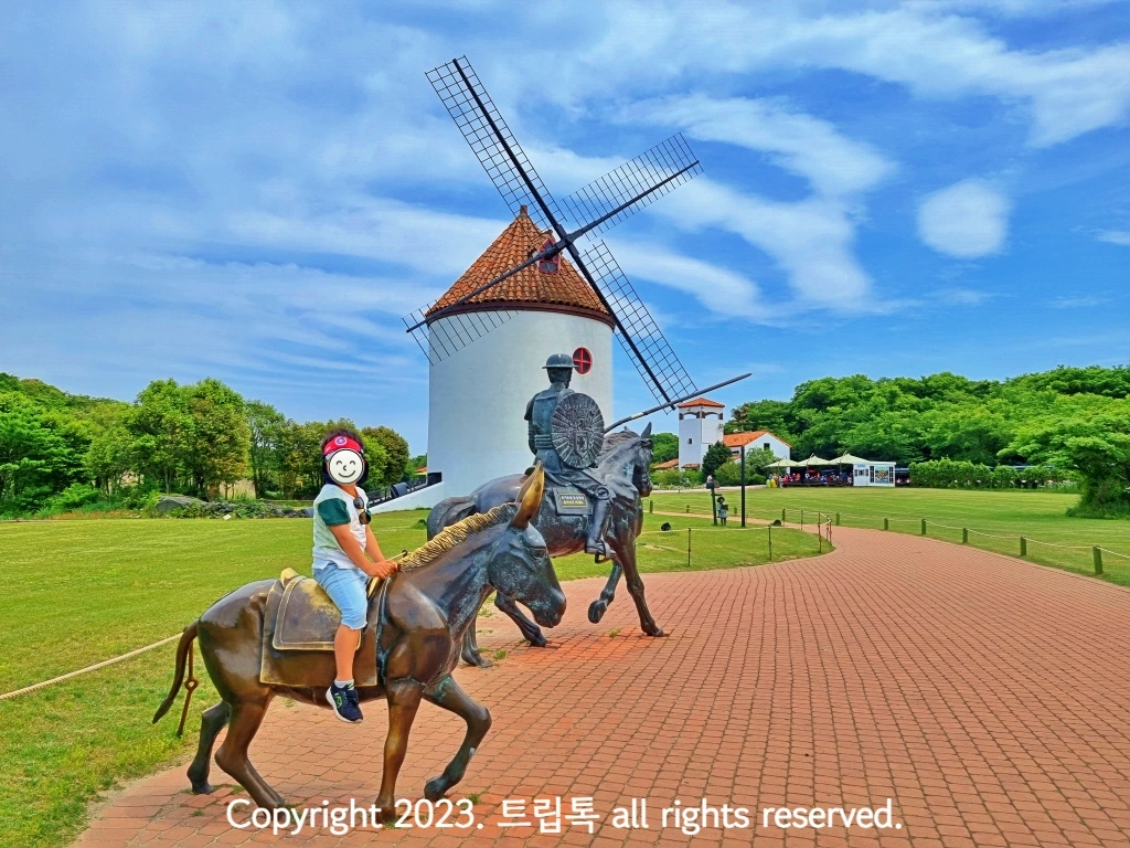 뒷편에 풍차가 있고 그 앞에 금속 당나귀를 탄 아이