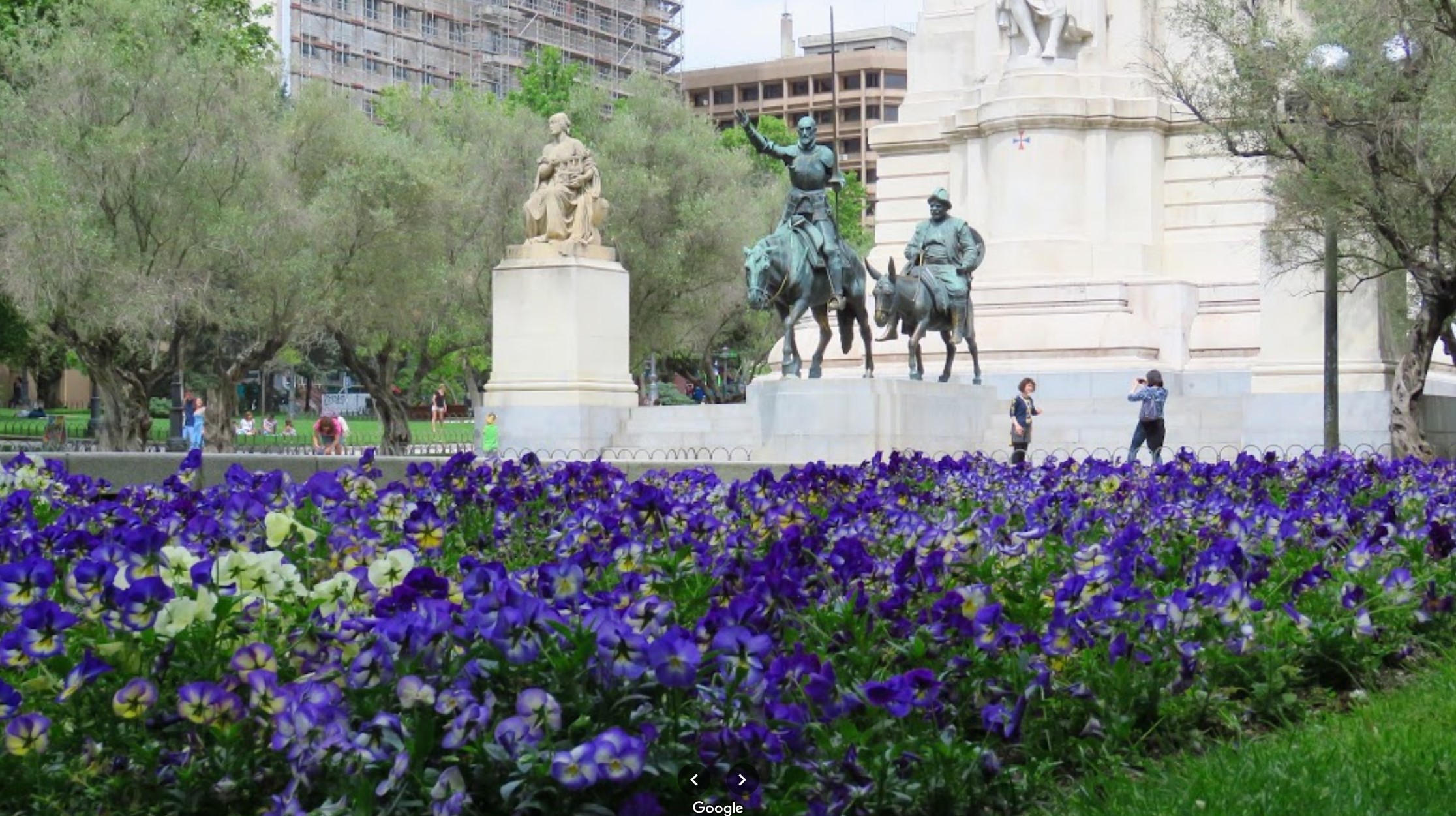 이미지에는 전경에 생생한 보라색과 흰색 꽃이 있는 아름다운 정원이 다채로운 디스플레이를 이루고 있는 모습이 나와 있습니다. 배경에는 스페인 마드리드 에스파냐 광장에 위치한 말을 탄 돈키호테와 산초 판차의 동상이 있다. 장면은 나무로 둘러싸여 있으며 몇몇 사람들이 평화로운 분위기를 즐기며 상징적인 기념물 근처에서 사진을 찍고 있는 모습이 보입니다. 전반적인 환경은 고요하며 자연과 역사적 예술이 조화를 이루고 있습니다.