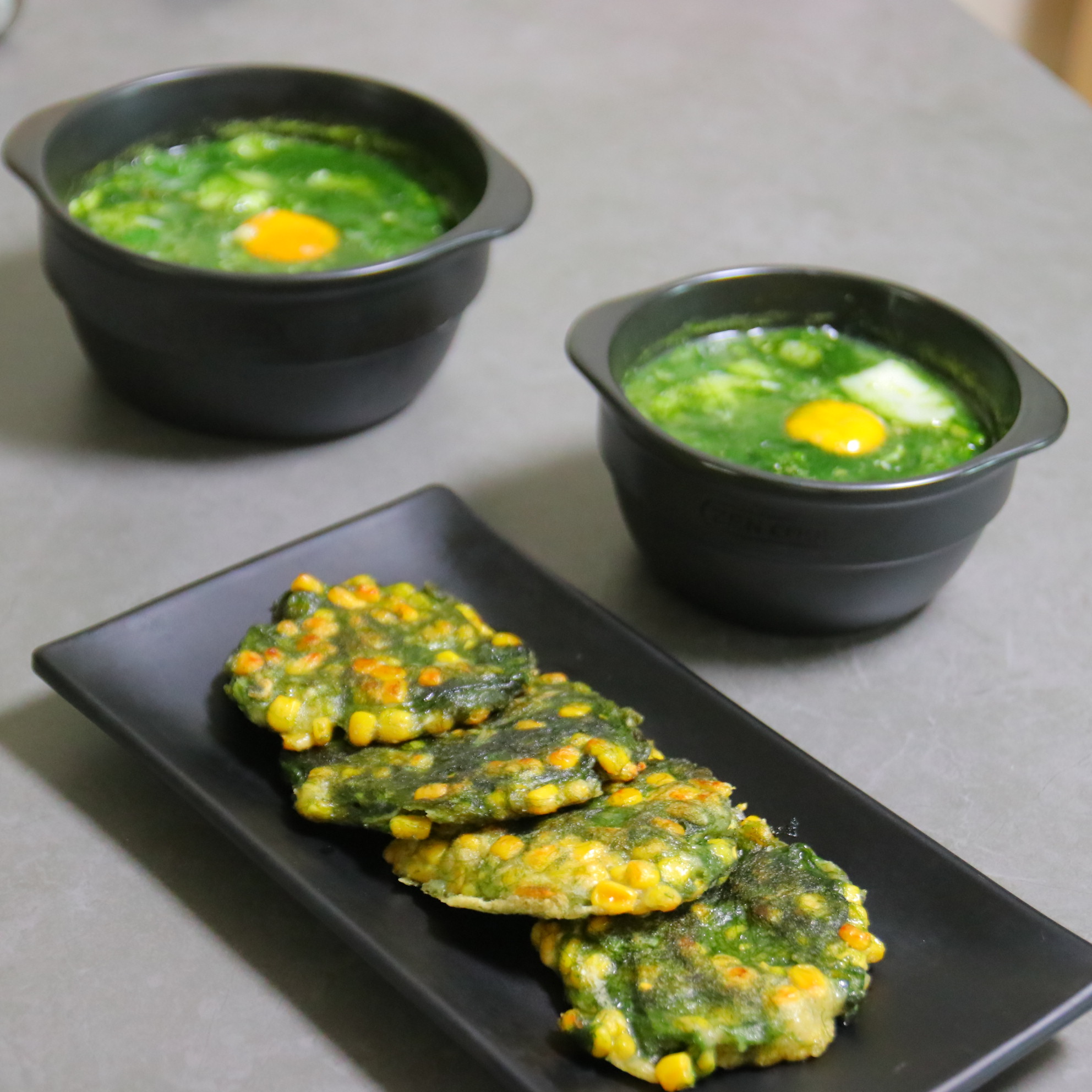 파래로 만든 죽을 검고 동그란 그릇에 가득 담아 놓고&#44; 그 밑에는 파래와 옥수수를 넣고 부친 파래전을 검고 직사각형의 접시에 4개 놓아두고 찍은 사진