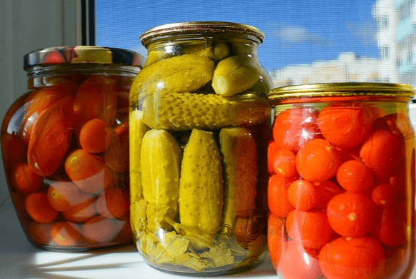 투명유리병에 통오이 피클이 가득 담겨있고 양옆에는 투명유리병에 빨간 야채 피클이 담겨있다.