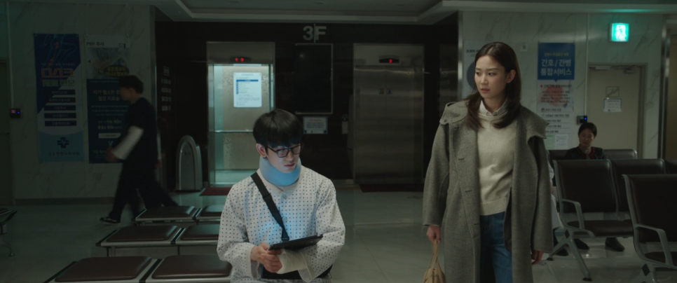 "병원 로비에서 환자복을 입은 남성과 코트를 입은 여성이 대화를 나누는 듯한 장면"