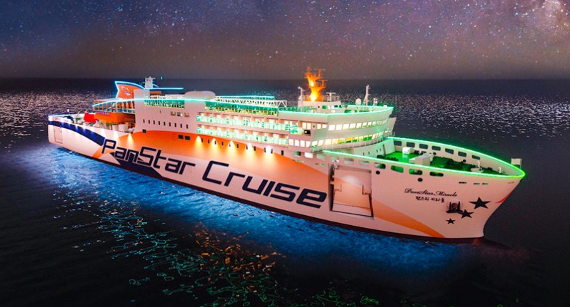 밤바다 위를 항해하는 팬스타미라클호 크루즈선. 선체에는 'PanStar Cruise'라는 대형 로고가 그려져 있으며, 선상에는 다양한 조명이 화려하게 빛나고 있다. 별이 가득한 밤하늘과 푸른 바닷물이 조화를 이루며 환상적인 분위기를 연출한다.