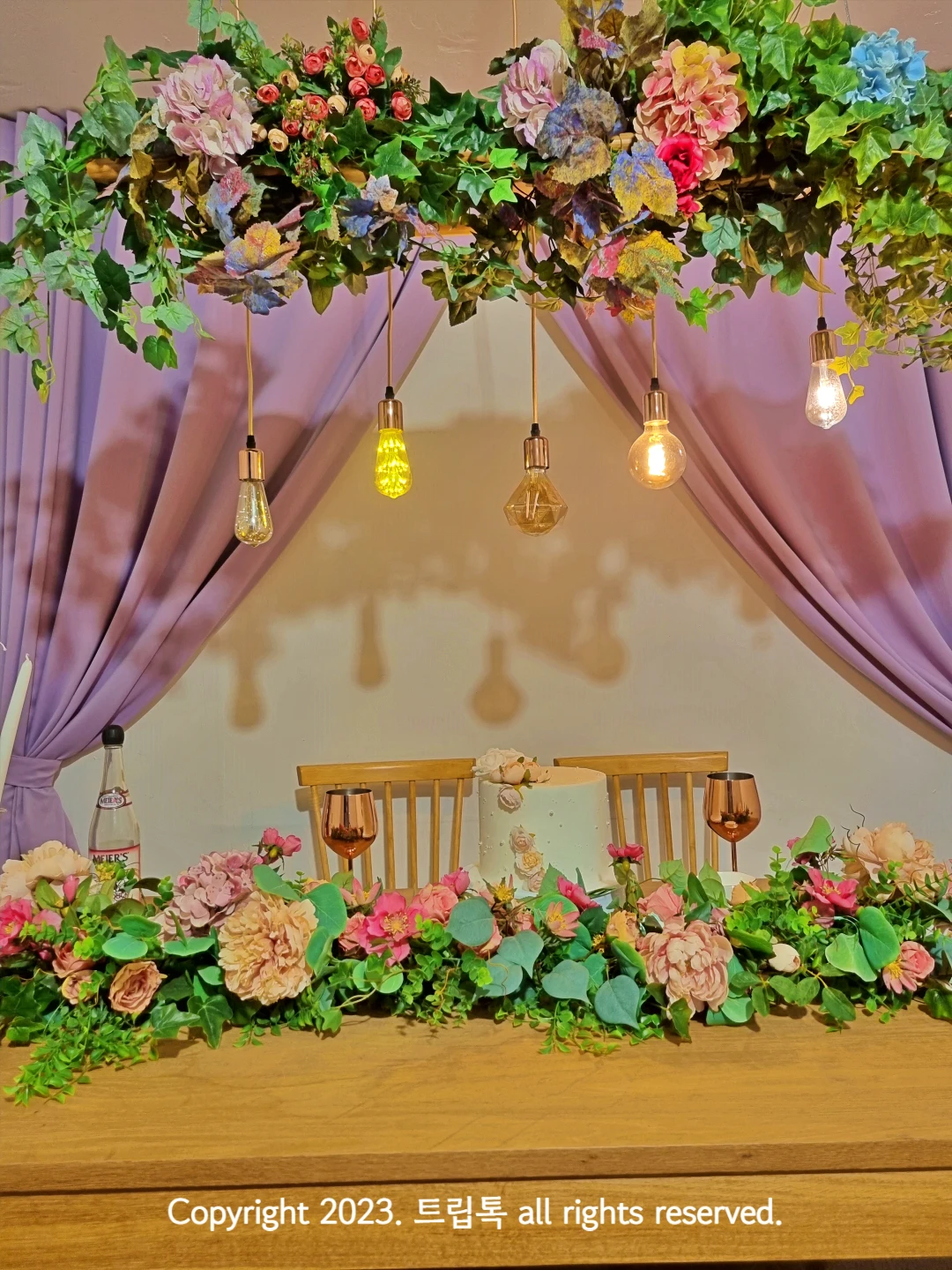 연보라색 커튼아래 테이블에 분홍색꽅장식이 있고 전구들이 천장에서 메달려있다
