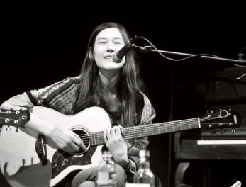 손지연이-기타를-치며-마이크-앞에-앉아-눈을-감고-미소를-지으며-노래를-부르고-있는-흑백-사진