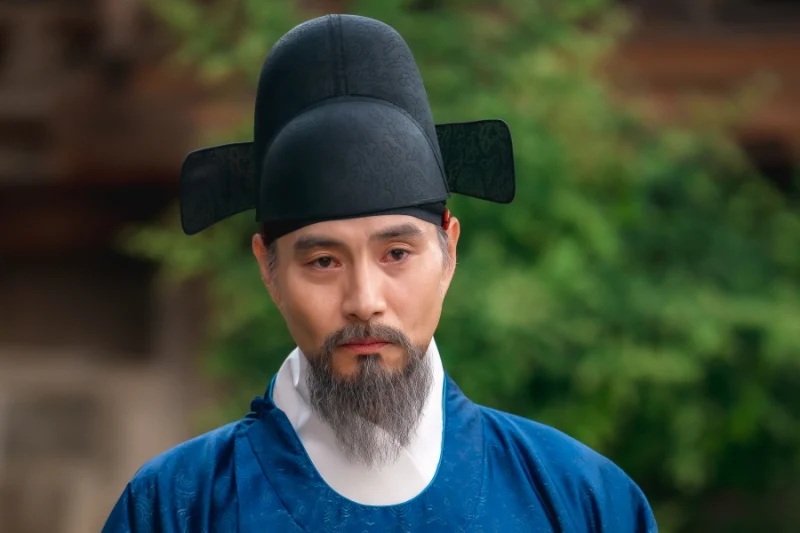 어느 실외를 배경으로 관직을 나타내는 갓에 파란 의복을 입고 서 있는 사극 드라마 혼례대첩의 조영배 캐릭터