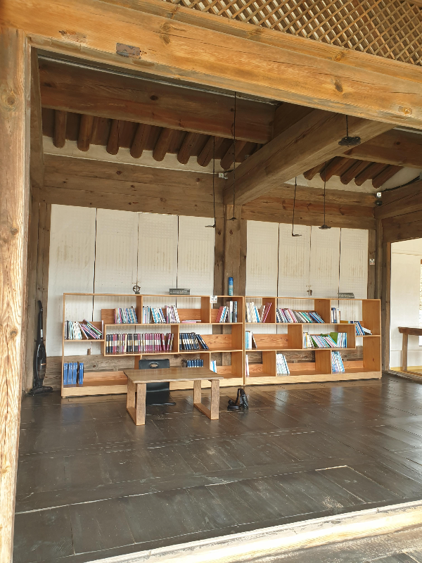 나무 마룻 바닥. 나무 기둥, 나무 들보로 되어 있는 책방. 뒤에 책장에는 책이 있다.