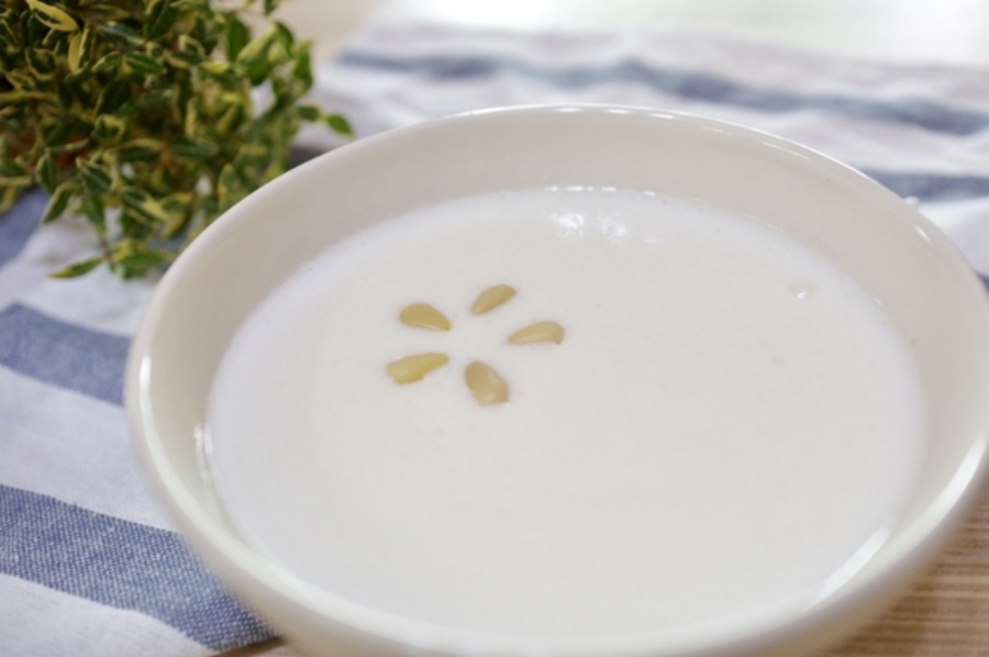 하얀 우유를 쌀과 함께 넣고 끓여서 만든 우유죽을 동그랗고 하얀 그릇에 가득 담아두고, 그 위에 잣을 5개 넣어두고 찍은 사진