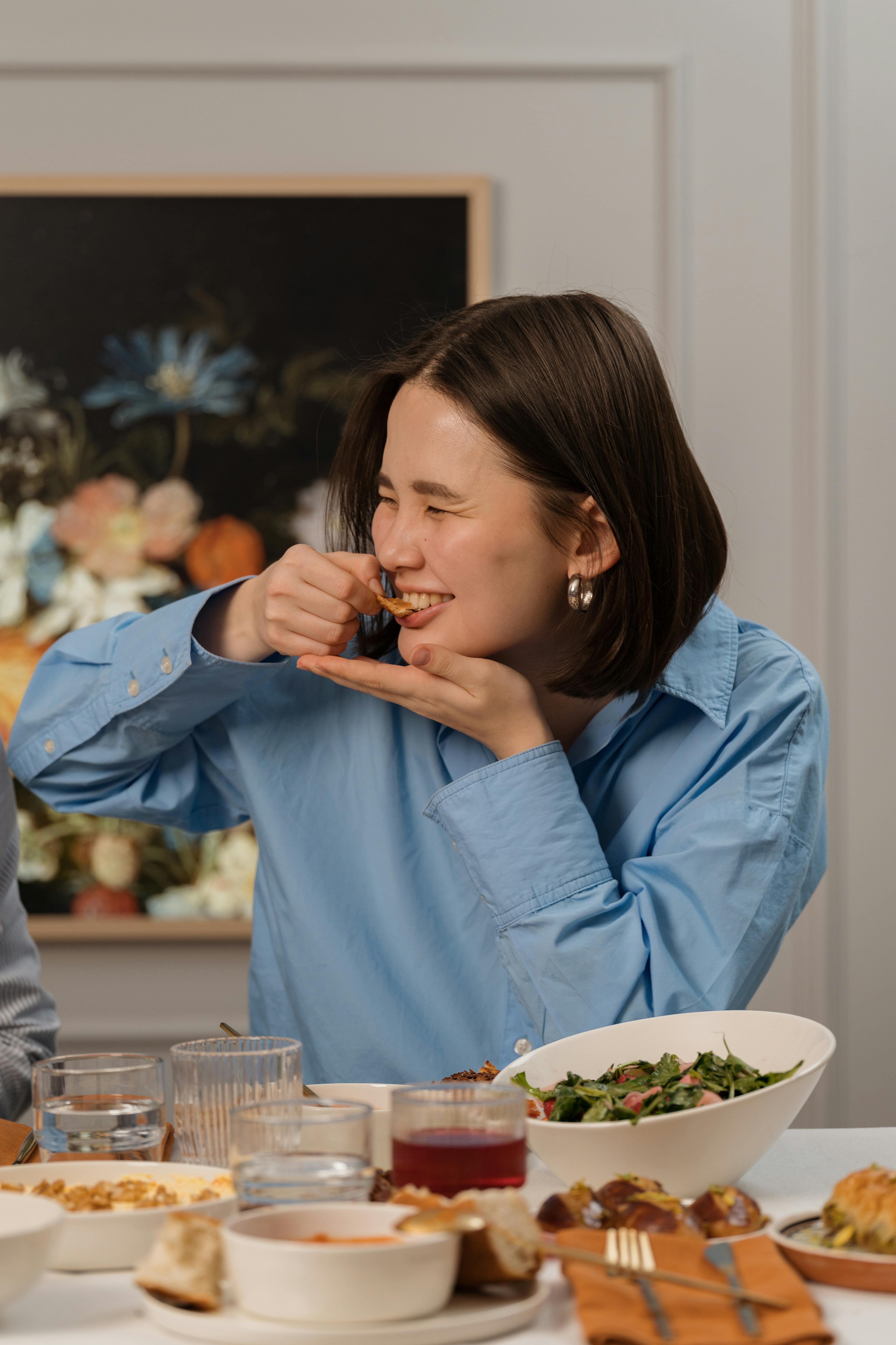 A young woman with short dark hair, wearing a light blue shirt and silver hoop earrings, is captured in a close-up shot as she enjoys a meal at a table set with various dishes, with a framed painting in the background