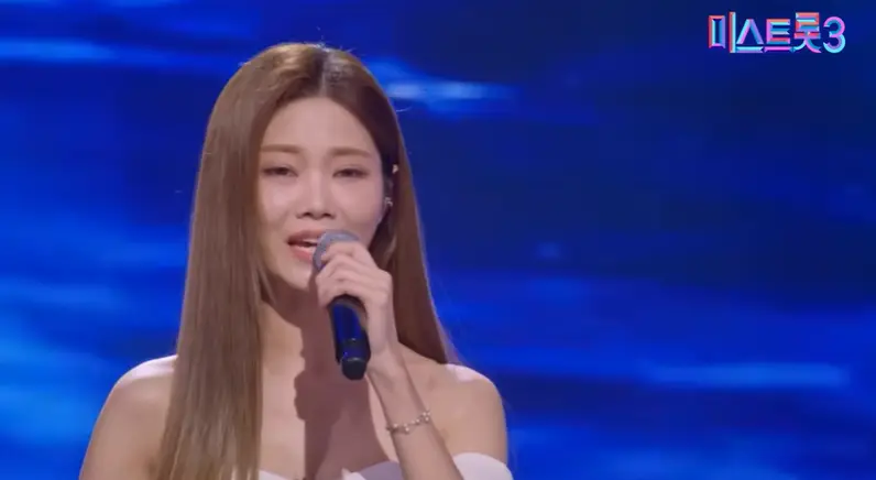 미스트롯3- 파란 무대배경 앞 흰 드레스 왼손으로 마이크를 들고 긴머리를 늘어뜨리고 노래하는 배아현