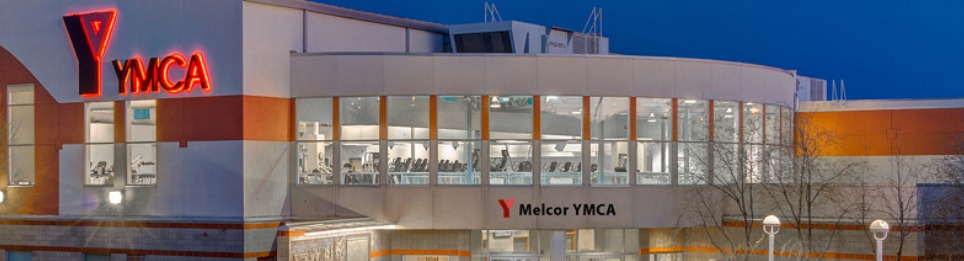 밤하늘 아래 불이 밝혀진 YMCA 건물이 보인다. 건물 외벽에는 빨간색 'YMCA' 로고가 선명하게 빛나고 있으며, 'Melcor YMCA'라는 이름도 함께 적혀 있다. 유리창 너머로는 실내 운동 기구들이 정렬되어 있고, 내부 조명이 따뜻하게 실내를 밝히고 있다.
