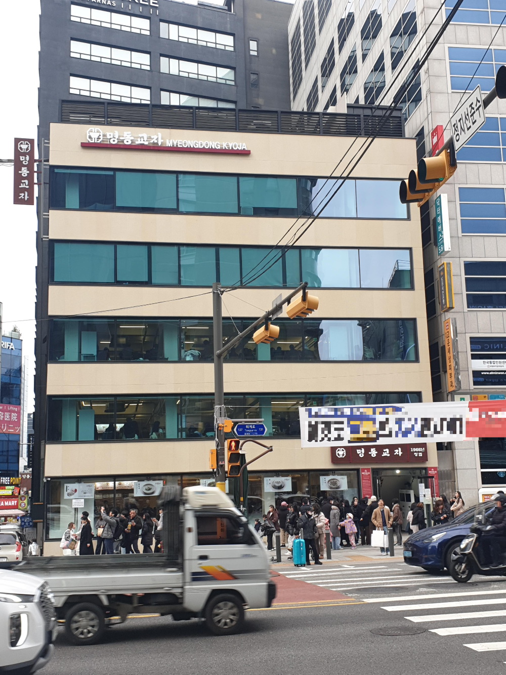 횡단보도 뒤 5층 명동교자 건물이 보인다. 건너편 횡단보도 앞에 사람들이 신호를 기다리고 있다.