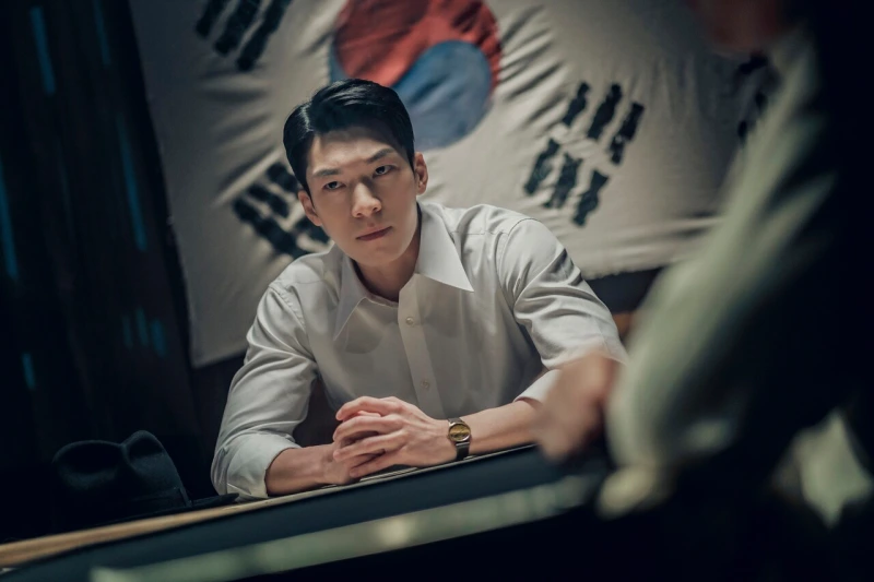 태극기를 배경으로 앉아 있는 넷플릭스 경성크리처에서 권준택을 연기하는 배우 위하준