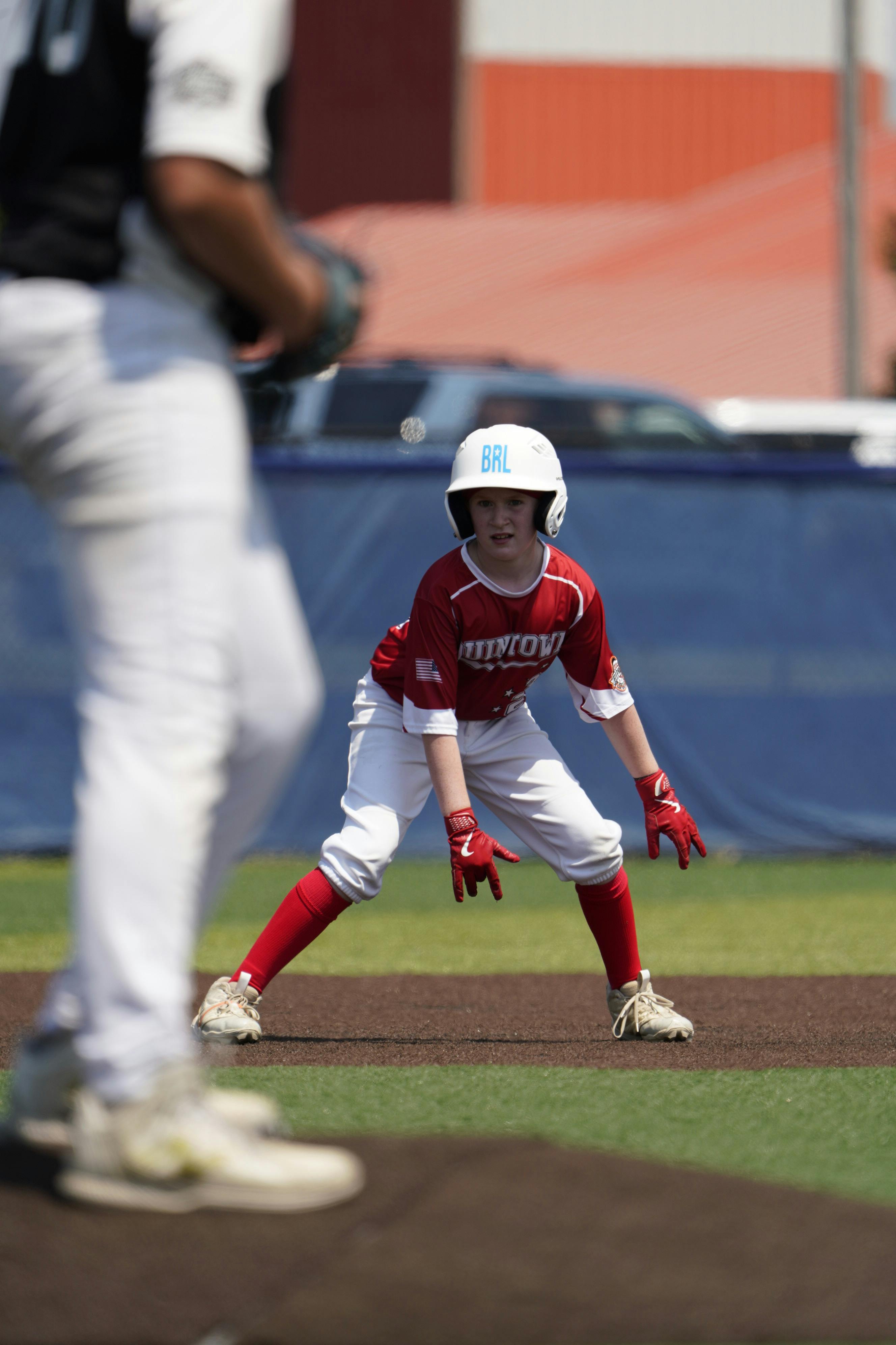 야구 선수가 1루 베이스 옆에서 주자 자세를 취하고 있는 모습. 카메라 초점은 앞쪽의 다른 선수에 맞춰져 있다.