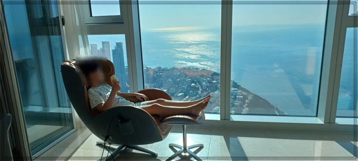 탁트인-바다전망이-보이는-높은-건물-숙소에서-바다를-바라보며-안마의자에-앉아있는-아이-아이스크림을-먹고-있음-환한-낮이며-반짝이는-오션뷰가-주된-경관이고-마을이-살짝-보임