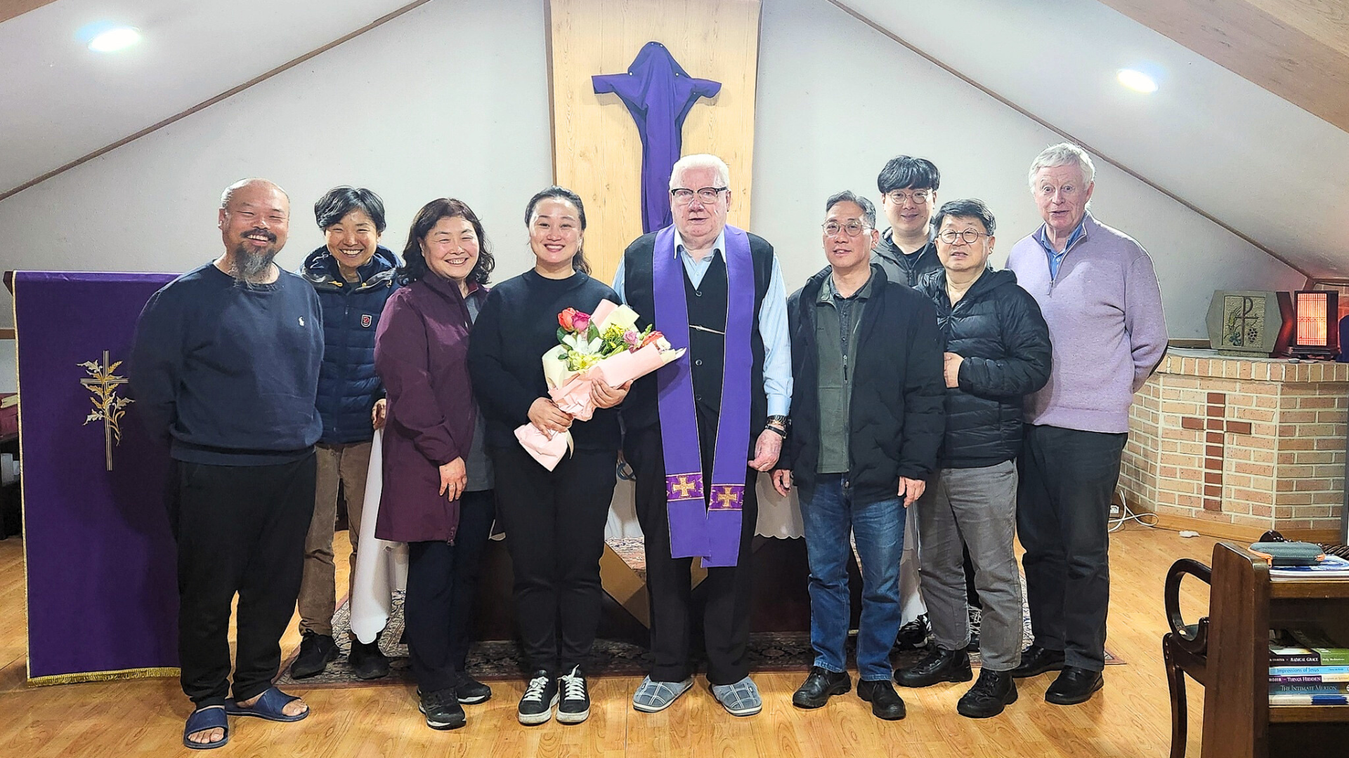 중앙에 커다란 나무 십자가가 있고 그 앞에 중앙에 교구님과 양쪽으로 여러 사람들이 일렬로 서서 앞을 바라보고 사진을 찍은 모습의 이미지