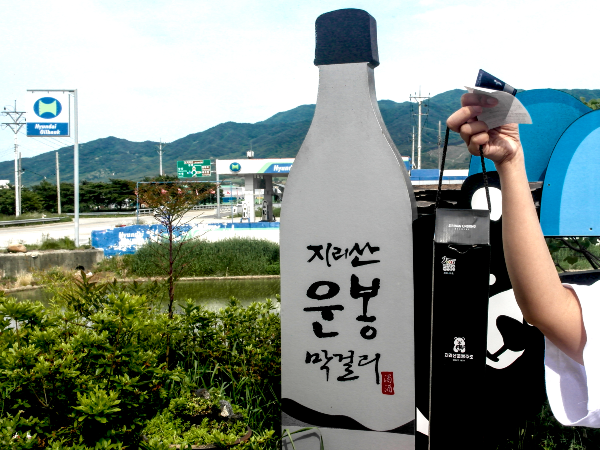 건물 앞에 있는 막걸리 모형에서 구매한 막걸리를 들고 한 컷