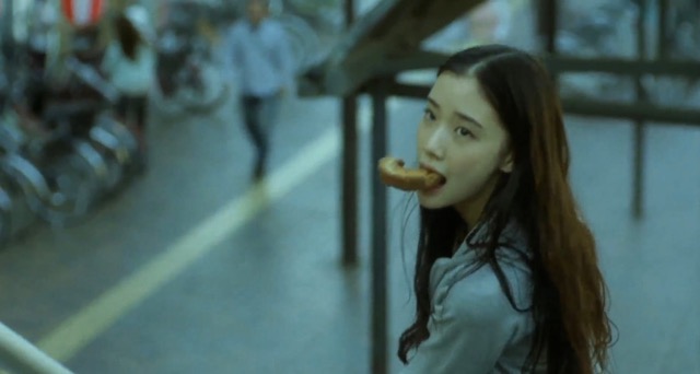 영화 《백만엔걸 스즈코》에서 스즈코가 도넛을 물고 무심한 듯 뒤를 돌아보는 장면. 평범해 보이는 일상 속에도 무언가를 벗어나고자 하는 자유의 갈망이 조용히 번진다.