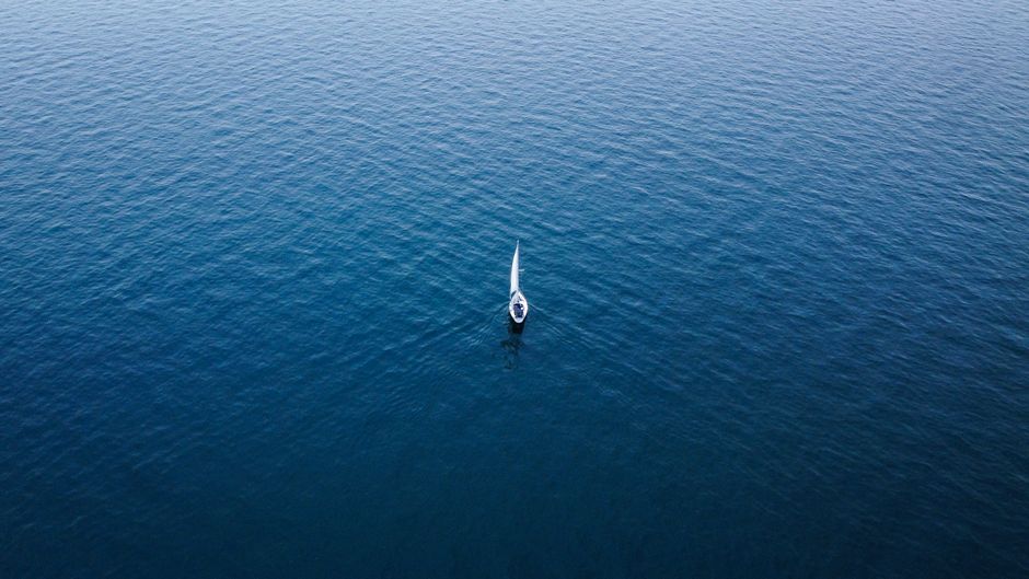 A single sailboat navigating calm blue waters from an aerial perspective, symbolizing solitude and adventure.