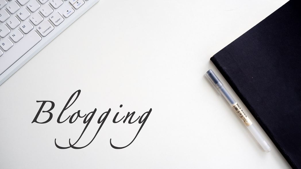 하얀 책상 위에 키보드와 노트 그리고 펜이 올려져 있다. 그리고 좌측 하단에 영어로 Blogging이라는 문구가 들어가 있다.