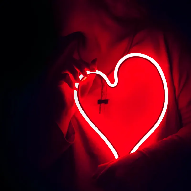 불이 꺼진 깜깜한 방에서 한 여성이 하트 모양의 붉은색 조명을 안고 있다.