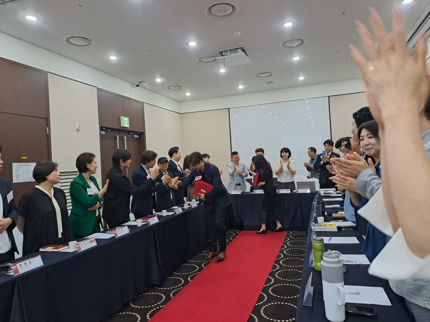 ㄷ자 회의실에 일어서서 30명의 멤버들과 하이파이브를 하며 자유온뷰티 스킨케어 김소영대표님과 룸프스튜디오 문민대표님이 손벽을 치며 손을 마주치고 있습니다. bni 그랜드챕터 가입을 환영합니다.