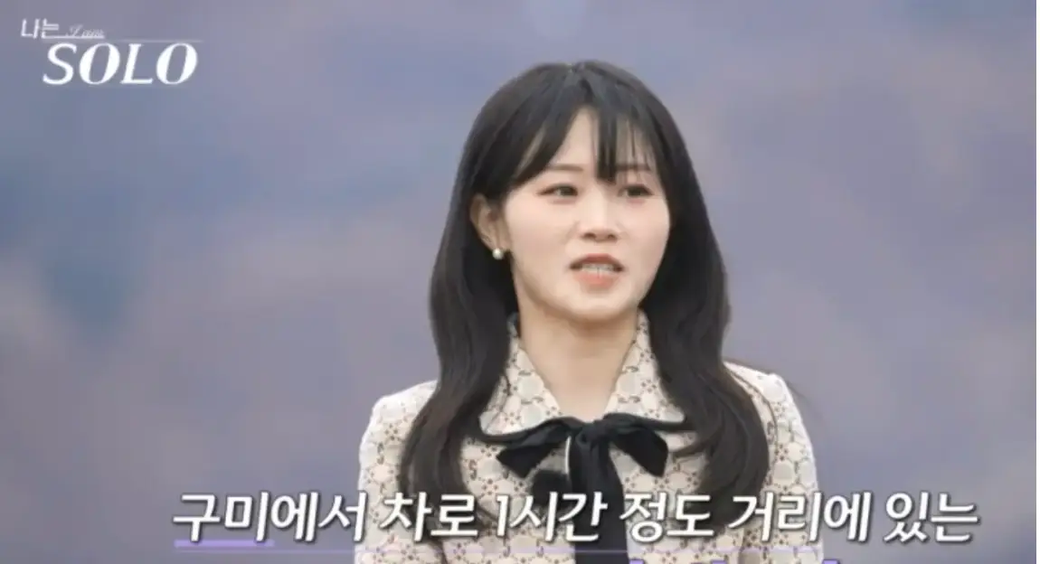 긴웨이브 머리 아이보리색 원피스 목에 검은 리본이 달린 옷을 입고 말하는 현숙 아래 흰글씨 구미에서 차로 1시간 정도 거리에 있는