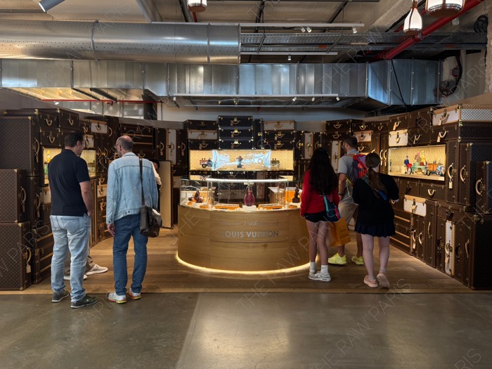 Visitors exploring the chocolate shop boutique at LV DREAM Louis Vuitton in Paris surrounded by iconic monogram trunks ❘ Des visiteurs découvrant la boutique de chocolats chez LV DREAM Louis Vuitton à Paris entourée de malles monogrammes