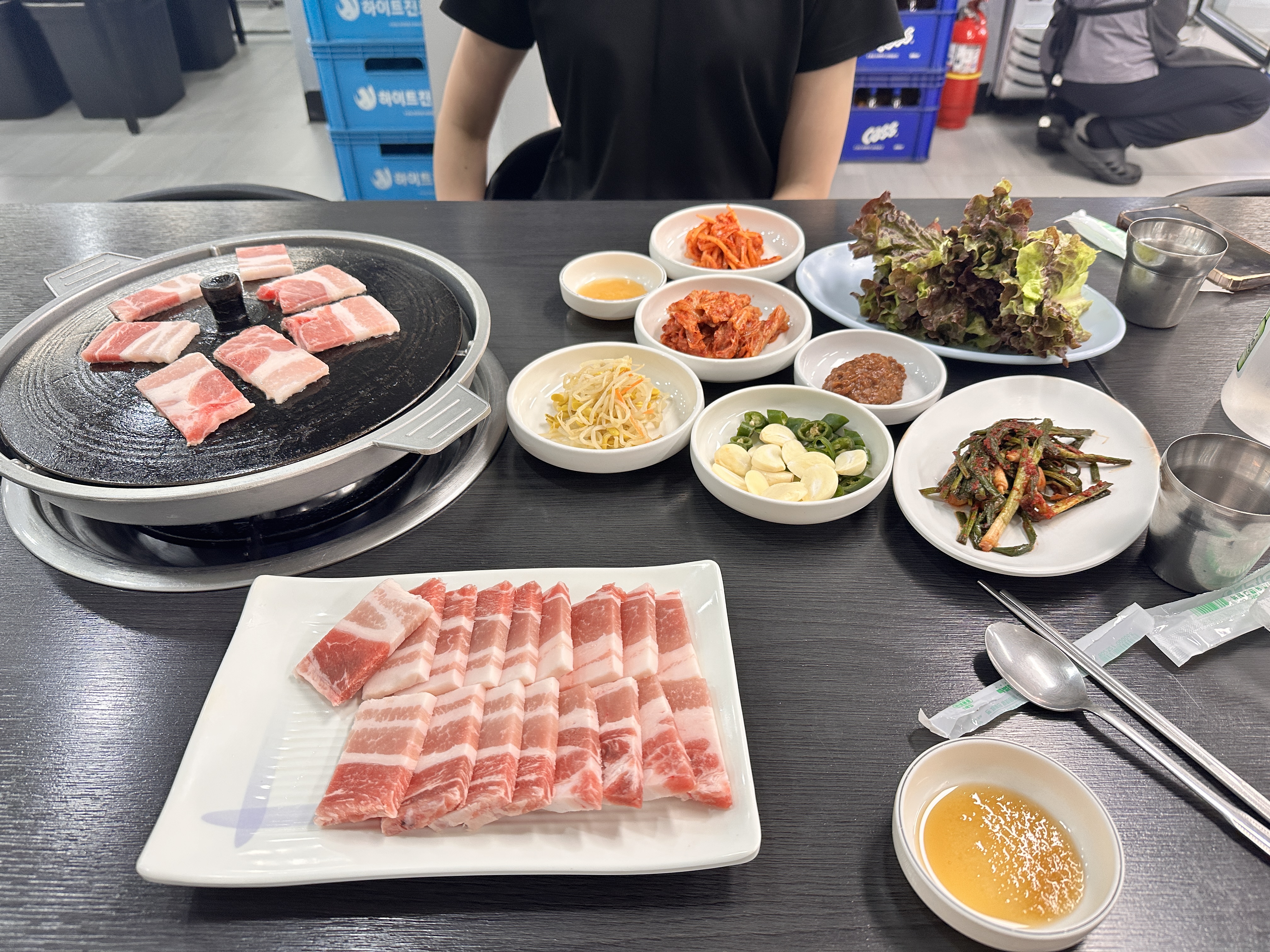 테이블 위에 솥뚜껑 불판과 냉동 삼겹살, 각종 한정식 밑반찬(콩나물, 김치, 쌈채소 등)이 차려진 전체 상차림