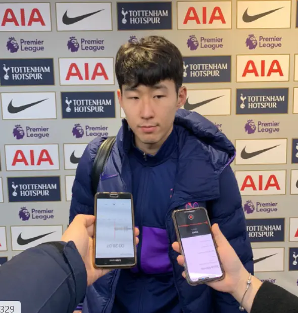 손흥민- 토트넘 광고벽보 앞 파란색 잠바를 입고 상대방을 바라보는 손흥민과 손흥민 앞으로 핸드폰을 들고 내밀어진 두손