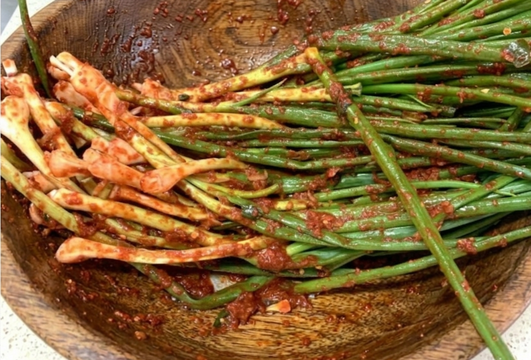 질감이 살아있는 나무 보울에 담긴 싱싱한 쪽파와 진하고 먹음직스러운 붉은색 파김치 양념.