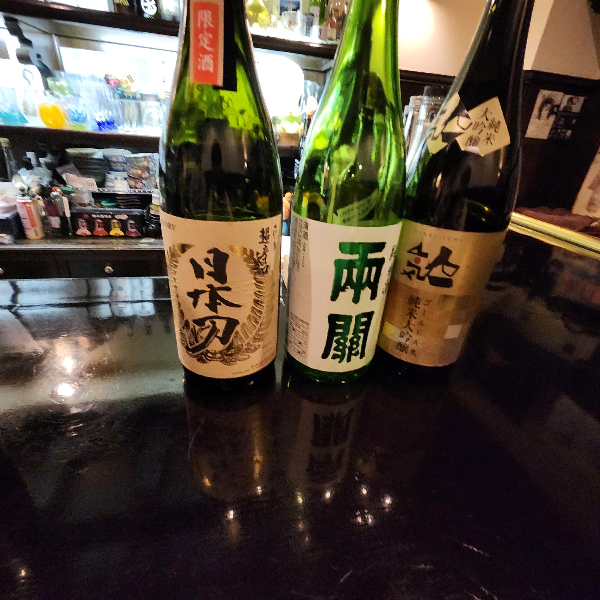 바 테이블 위에 사케 3병이 올려져 있다. 니혼토, 료칸, 스즈키라고 쓰여 있다.