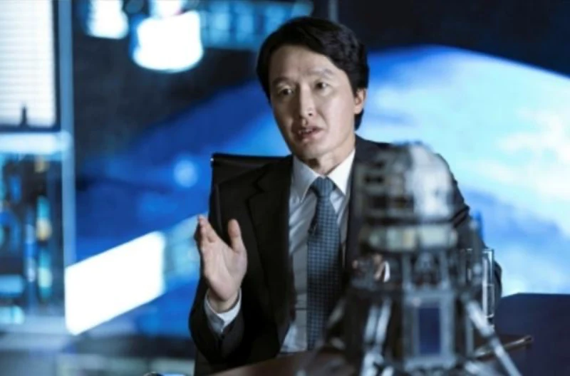 어느 사무실을 배경으로 검은 정장을 입고 의자에 앉아 있는 영화 더문의 오규석