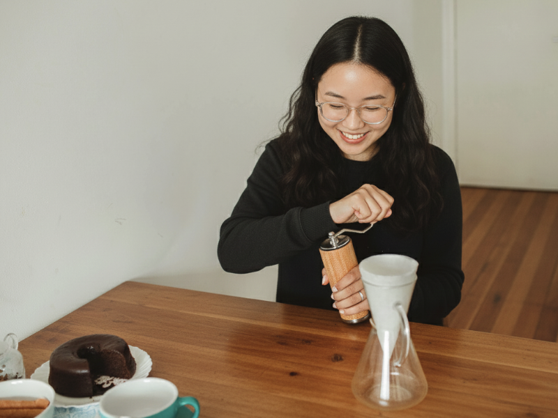 목재 테이블 위에 케멕스 3컵과 케익이 올려져 있다. 그리고 의자에 앉아 원두를 그라인딩 중인 사람