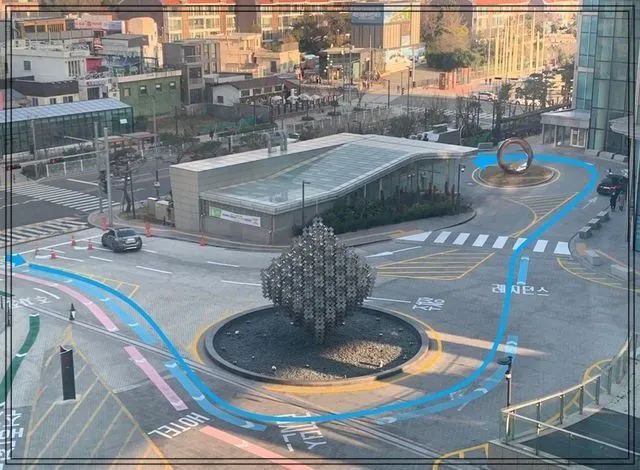 가운데-큐브모양의-조형물이-있고-이를-둘러싸고-파란색-주차유도선과-분홍색-유도선이-있음-엘시티-레지던스-주차장으로-들어가는-주차-유도선을-보여주는-모습-멀리-건물들이-보임
