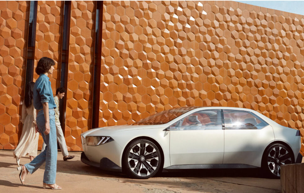 BMW Neue Klasse 전기차가 주황빛의 기하학적 패턴이 돋보이는 건축물 옆에 주차되어 있습니다. 보행자들이 차량의 혁신적인 디자인을 감상하고 있습니다