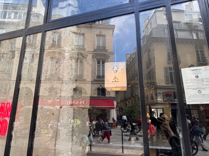 The stylish glass storefront window of SAMSO Korean noodle restaurant in Paris reflecting historic European buildings ❘ La vitrine du restaurant de nouilles coréennes SAMSO à Paris