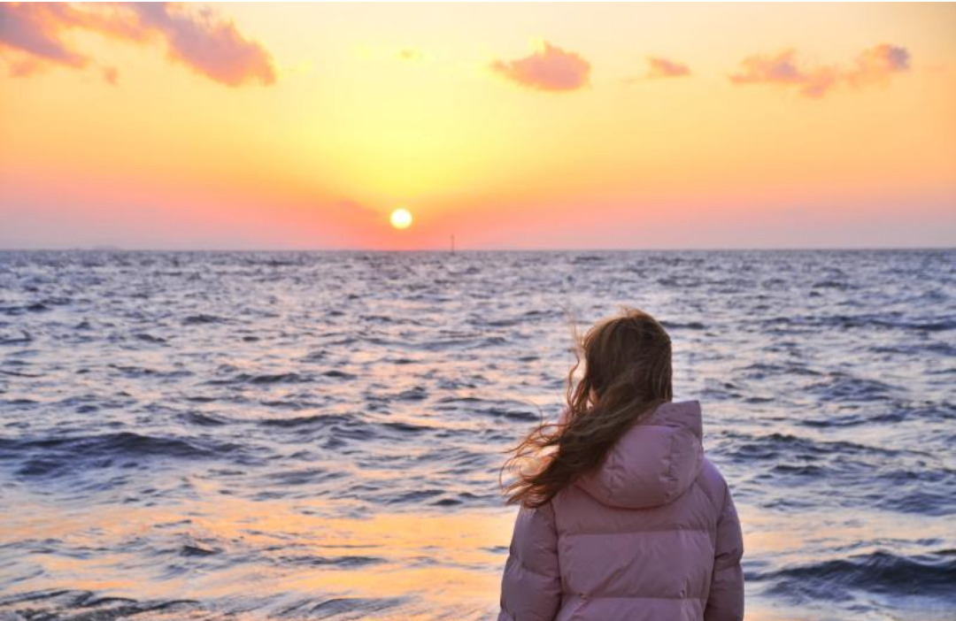 핑크 패딩을 입은 여자가 해변가에서 일출을 보고 있다.