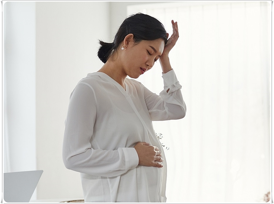 한손으로는 배를 가만히 감싸고 한 손으로는 이마에 가져다댄 채 다소 힘든 표정을 짓고 있는 임산부의 모습