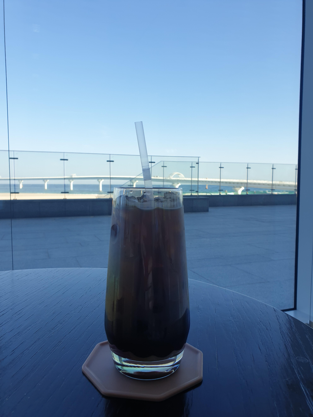 창 밖으로 바다가 보인다. 테이블 위에는 커피가 가득찬 빨대가 꽃힌 유리컵이 있다.