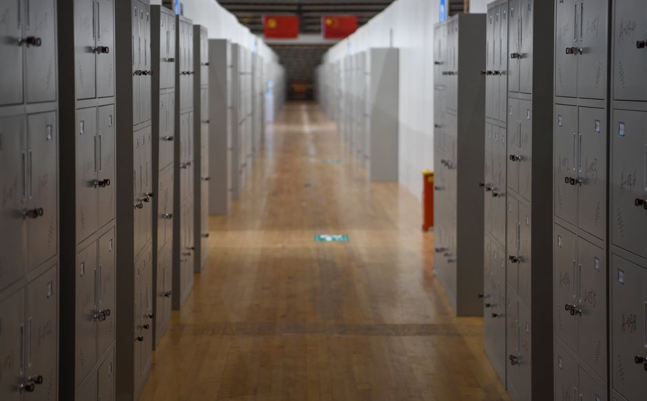A perspective view of a long hallway featuring rows of metal lockers and wooden flooring.