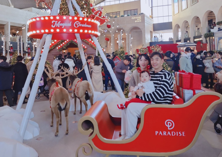 인천 파라다이스 시티 크리스마스 트리 앞 썰매 포토존에서 가족이 함께 사진 촬영하는 모습. 산타 썰매와 루돌프 장식이 있는 크리스마스 포토존 분위기